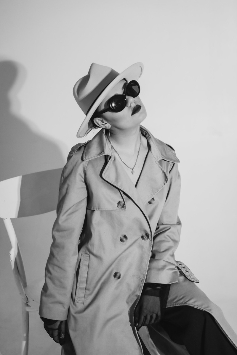 A woman in a trench coat, hat and sunglasses, black and white.